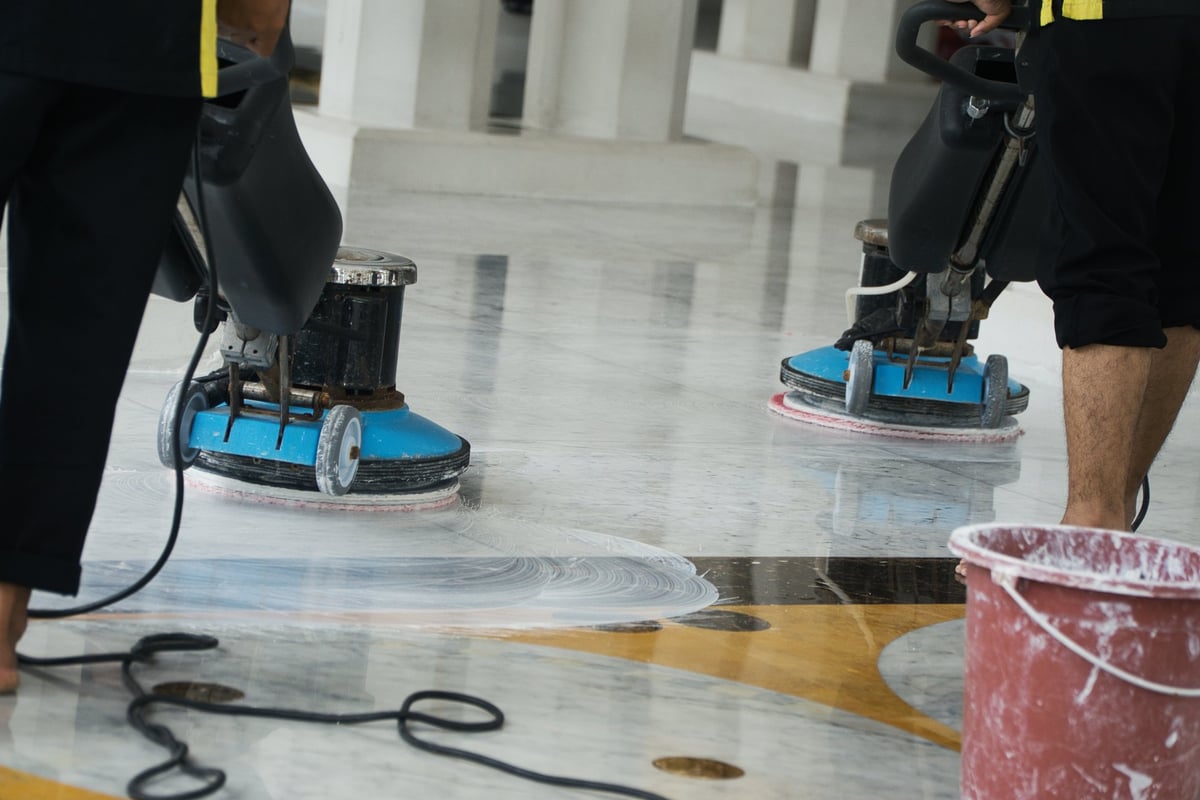 Two workers operate buffing machines to thoroughly clean a polished marble floor, ensuring it maintains its professional and spotless appearance